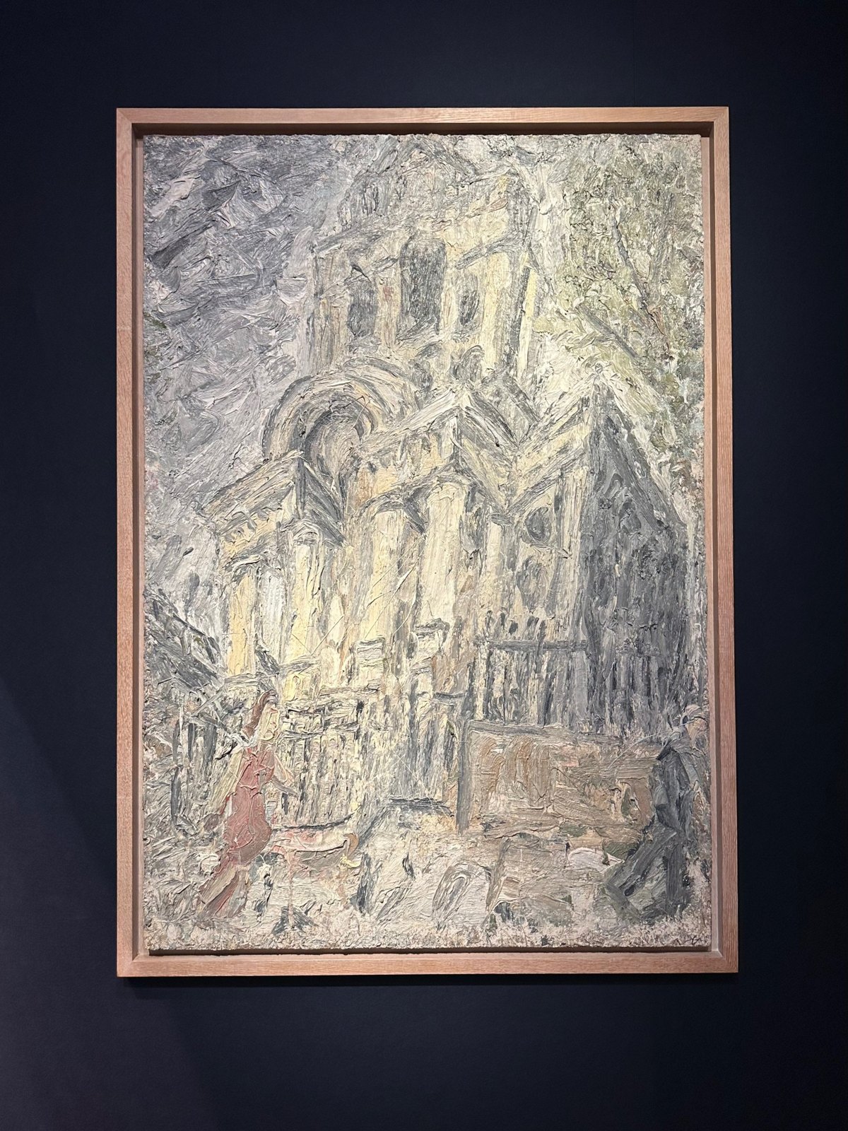 A thickly painted oil of a church in pale yellow, grey and green, in a pale oak frame on a dark navy wall